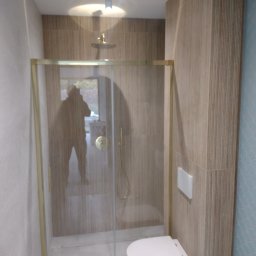 Remonty wnętrz mieszkań i domów Mariusz Ura - Nowoczesna kabina prysznicowa z złotymi ramami, zamontowana toaleta podwieszana, jasne płytki imitujące drewno na ścianach, niebieska ściana z wzorem rybiej łuski w tle.