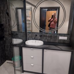 Remonty wnętrz mieszkań i domów Mariusz Ura - Nowoczesna łazienka z kabiną prysznicową typu walk-in, umywalką nablatową na szafce z szufladami i okrągłym lustrem, w którym odbija się osoba robiąca zdjęcie.