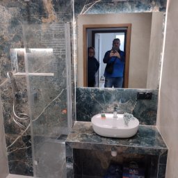 Remonty wnętrz mieszkań i domów Mariusz Ura - Wykończona łazienka z kabiną prysznicową i umywalką na blacie z płytek imitujących marmur. W lustrze odbija się osoba robiąca zdjęcie.