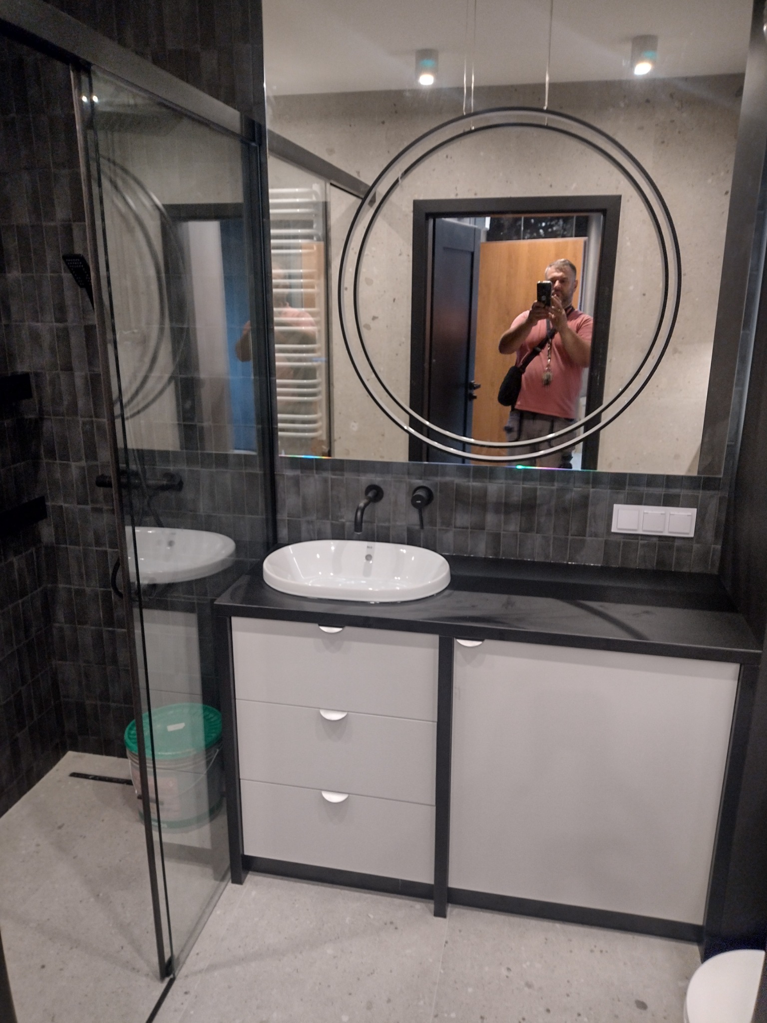 Nowoczesna łazienka z kabiną prysznicową typu walk-in, umywalką nablatową na szafce z szufladami i okrągłym lustrem, w którym odbija się osoba robiąca zdjęcie.