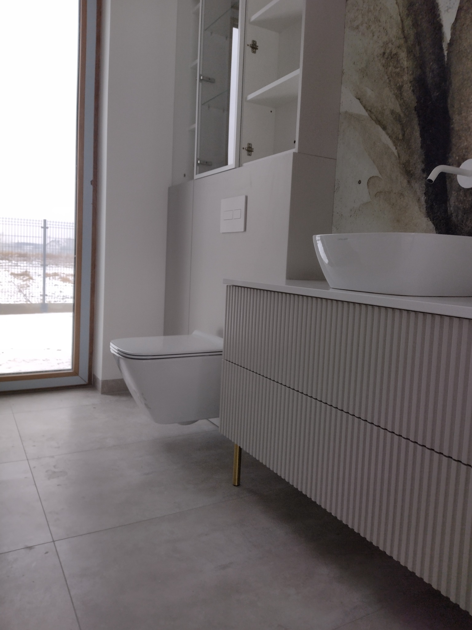 Nowoczesna łazienka w minimalistycznym stylu: podwieszana toaleta, umywalka nablatowa Catalano na szafce z ryflowanym frontem, szafka wisząca z przeszkleniami, abstrakcyjna grafika na ścianie.