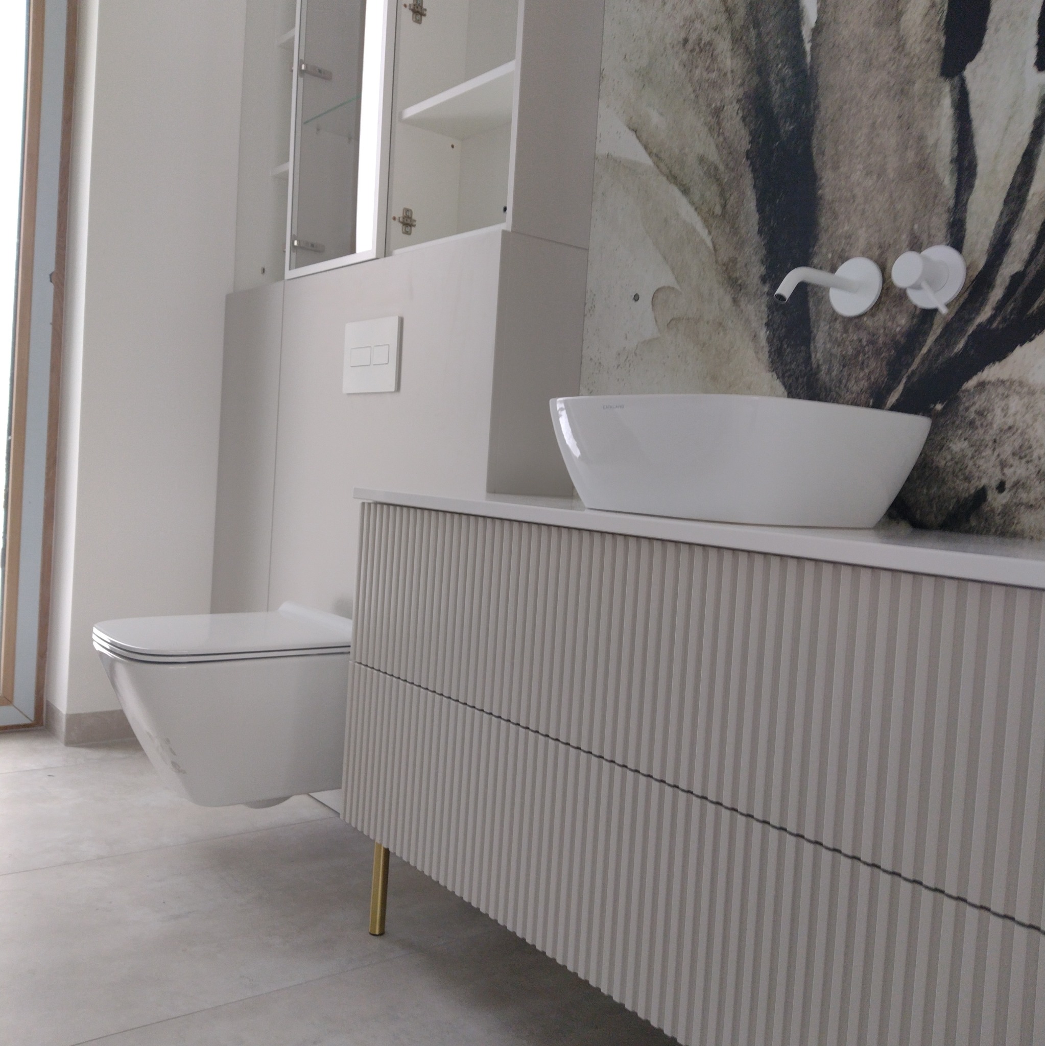 Nowoczesna łazienka w odcieniach bieli i szarości z wiszącą toaletą, umywalką nablatową Catalano oraz szafką z ryflowanym frontem na złotych nóżkach, na ścianie dekoracyjna tapeta.