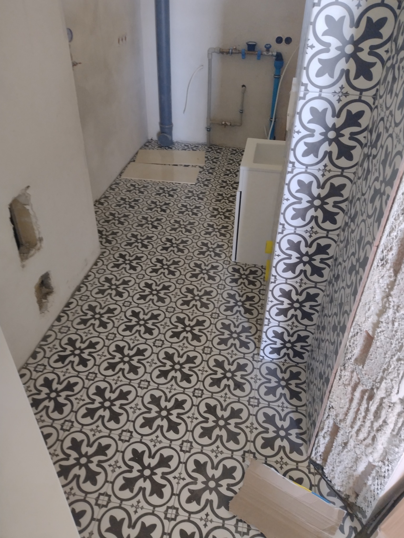Ukończona podłoga i fragment ściany z dekoracyjnymi, czarno-białymi płytkami w łazience. Widoczne instalacje wodne i fragment niezabezpieczonej ściany.