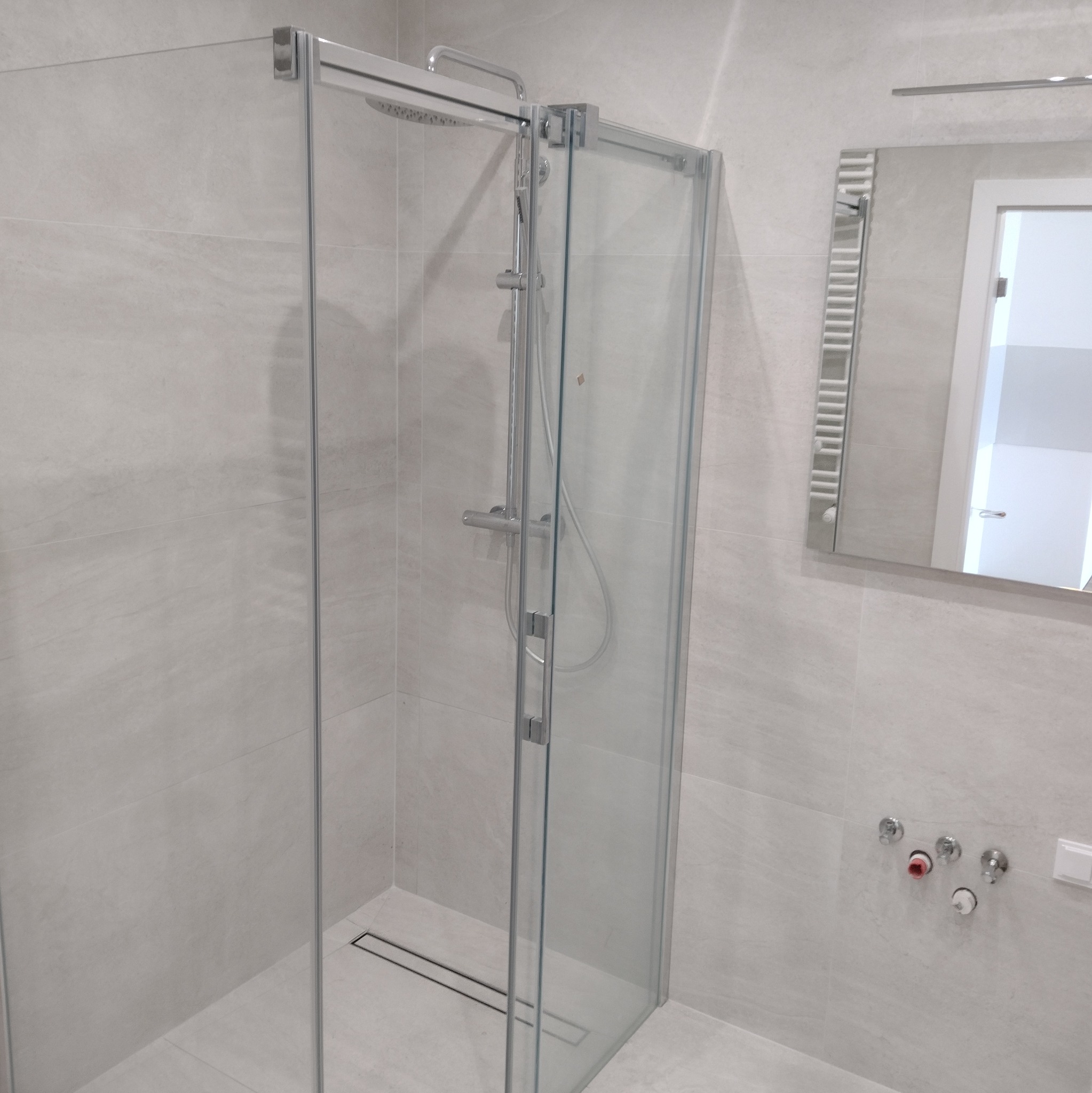 Nowoczesna kabina prysznicowa z odpływem liniowym, jasne płytki imitujące kamień, minimalistyczne baterie i lustro z grzejnikiem drabinkowym w tle.