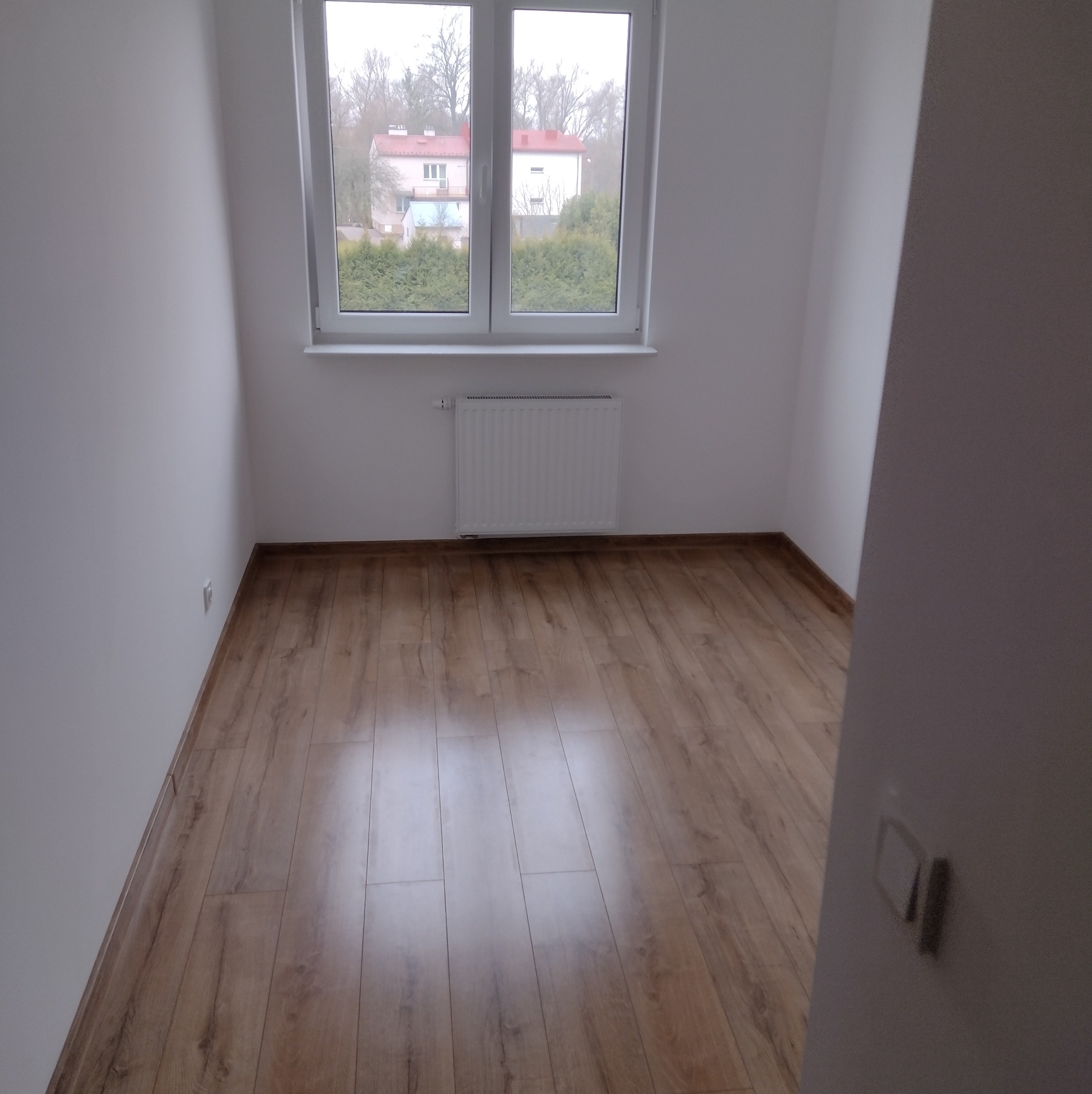 Jasne wnętrze pokoju po remoncie: panele podłogowe, białe ściany, okno z widokiem na zieleń. Minimalistyczny grzejnik pod oknem. Widoczny fragment włącznika światła.