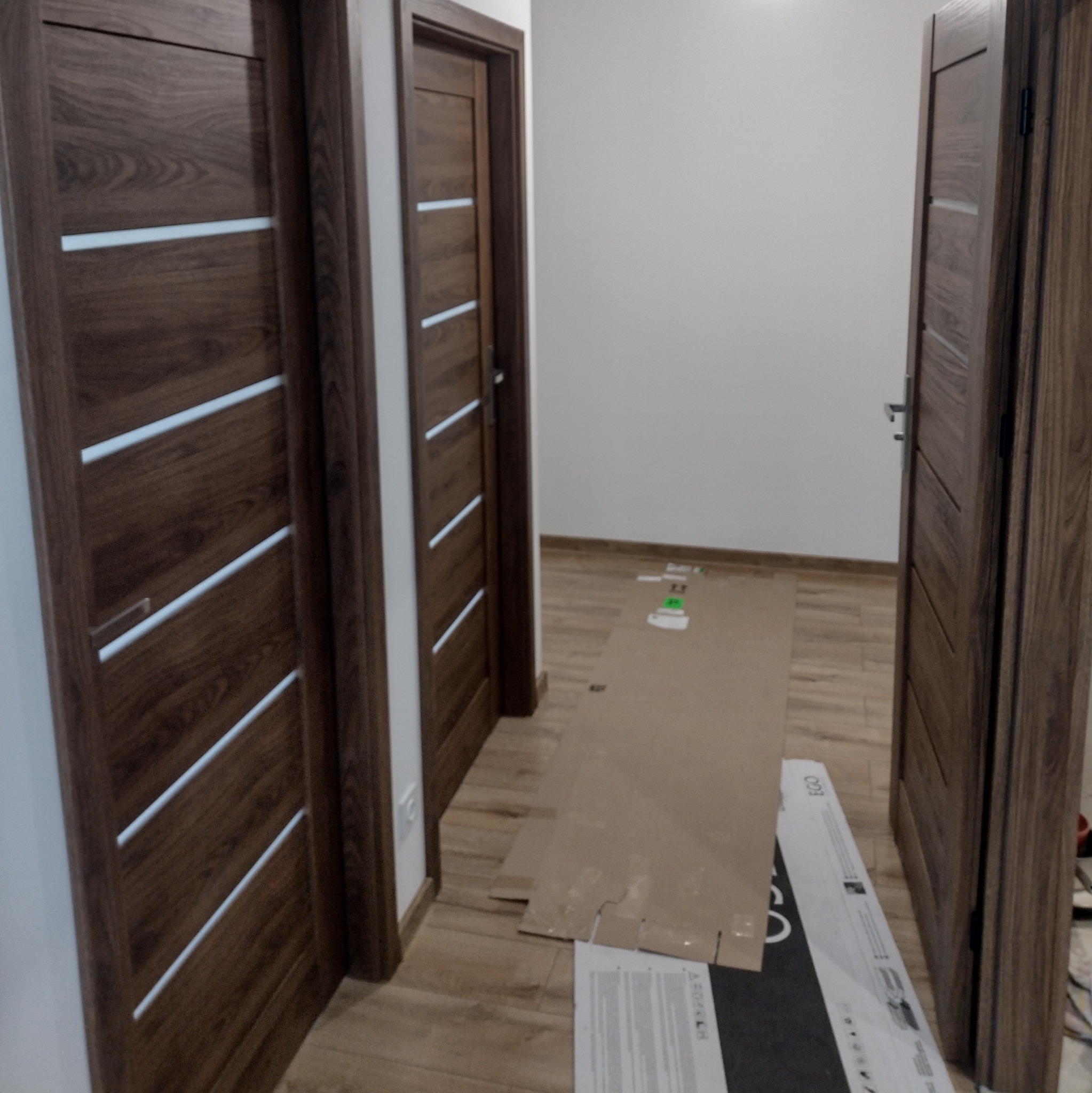 Nowoczesne, brązowe drzwi wewnętrzne z poziomymi, białymi wstawkami, zamontowane w jasnym korytarzu. Częściowo widoczne materiały zabezpieczające podłogę.