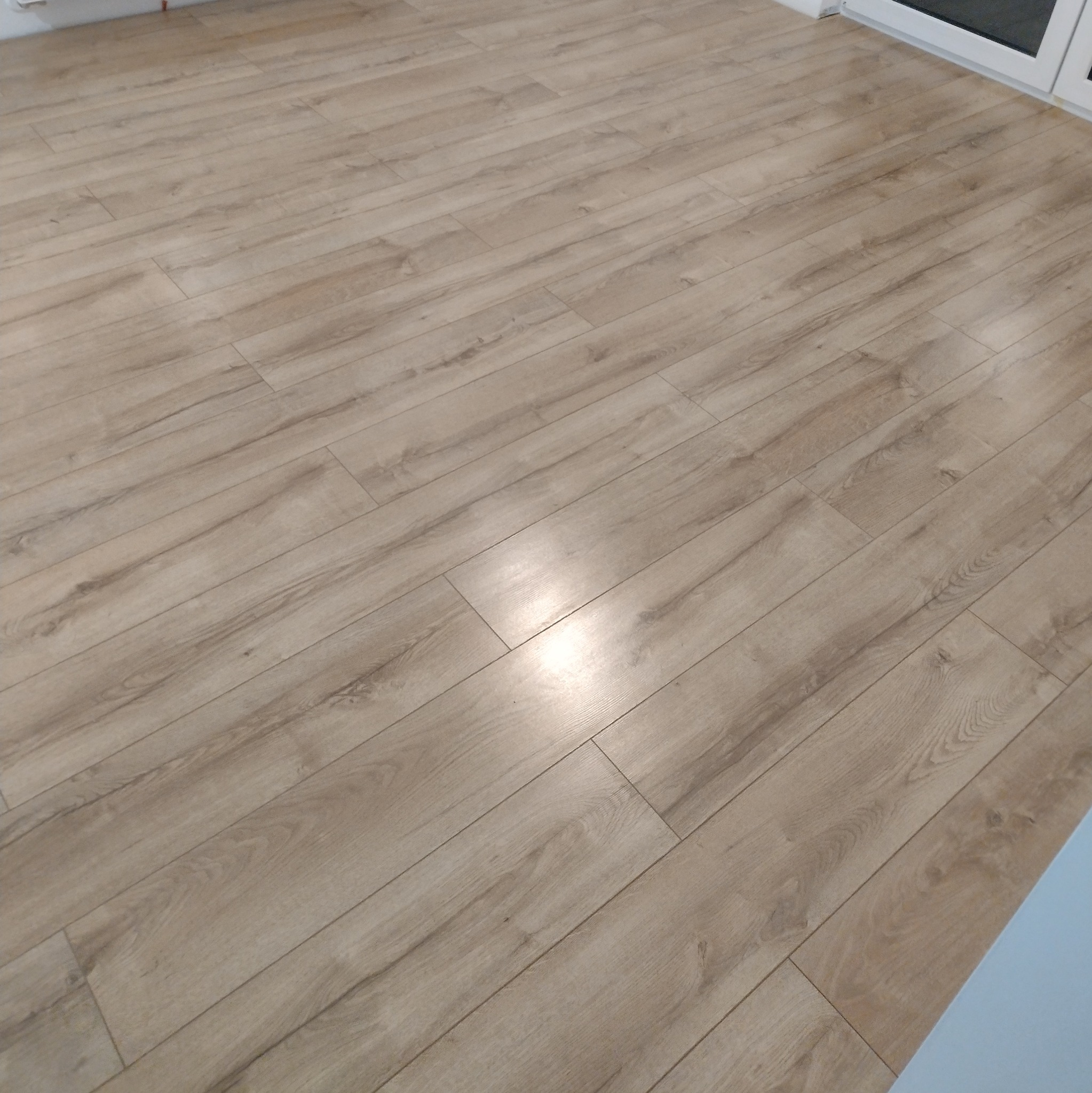 Jasne panele podłogowe ułożone w jodełkę, widoczne delikatne usłojenie drewna i subtelne refleksy światła. W tle fragment białej ramy okiennej.