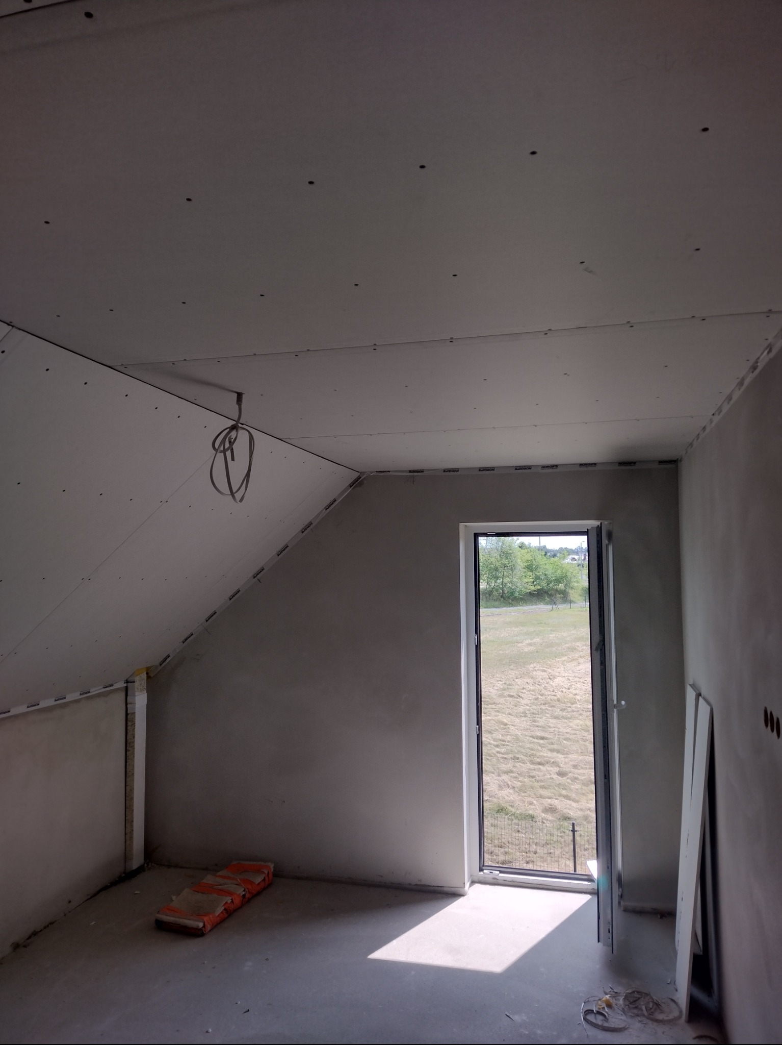 Poddasze w trakcie prac wykończeniowych: ściany i skośny sufit obłożone płytami karton-gips, widoczne łączenia, okno z widokiem na zewnątrz, przewody elektryczne.