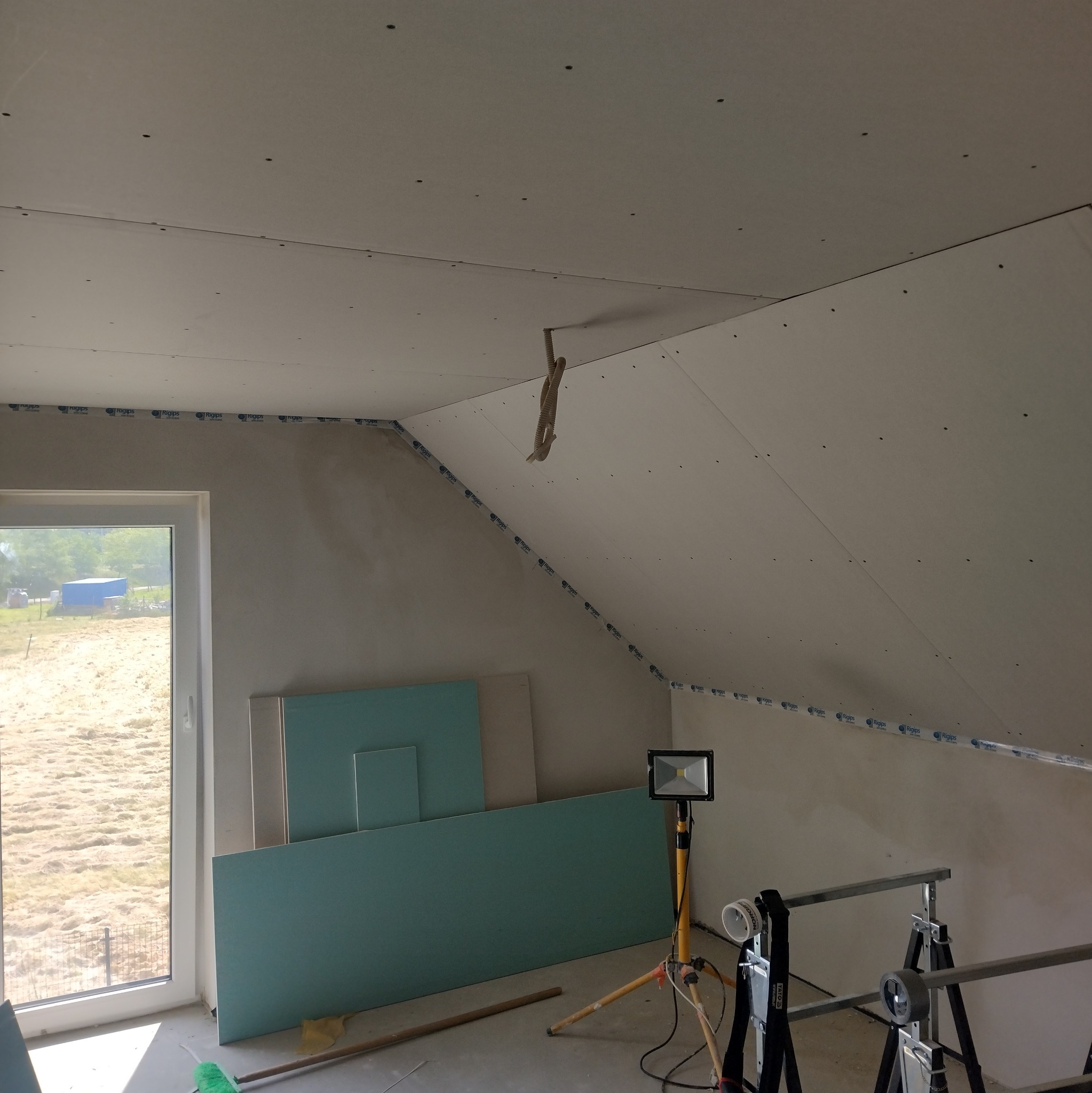 Skośne poddasze w trakcie prac wykończeniowych: zamontowane płyty gipsowe, widoczne narzędzia i oświetlenie budowlane. Okno z widokiem na zewnątrz.