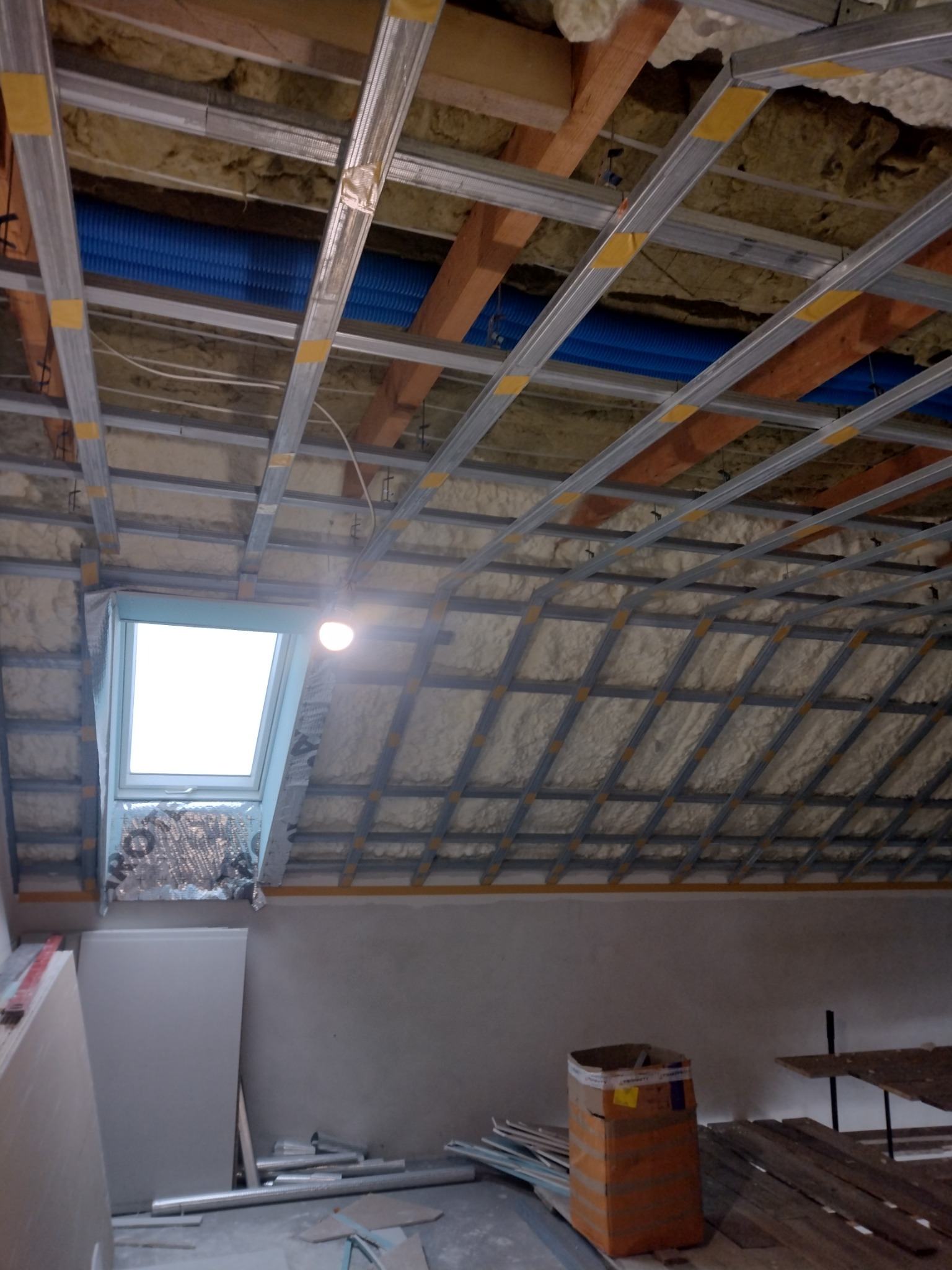 Skośny strych w trakcie izolacji: wełna mineralna między krokwiami, metalowy stelaż pod płyty gipsowe, okno dachowe. Widoczne materiały budowlane.
