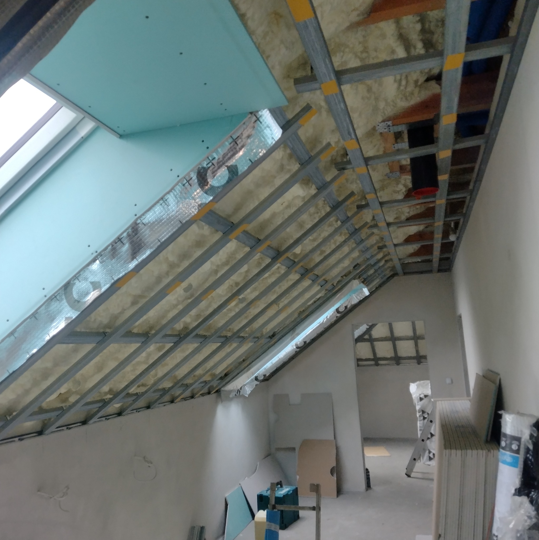 Montaż płyty G-K na poddaszu: widoczne profile, izolacja i częściowo zamontowane płyty. Prace wykończeniowe w trakcie realizacji, narzędzia i materiały budowlane w tle.