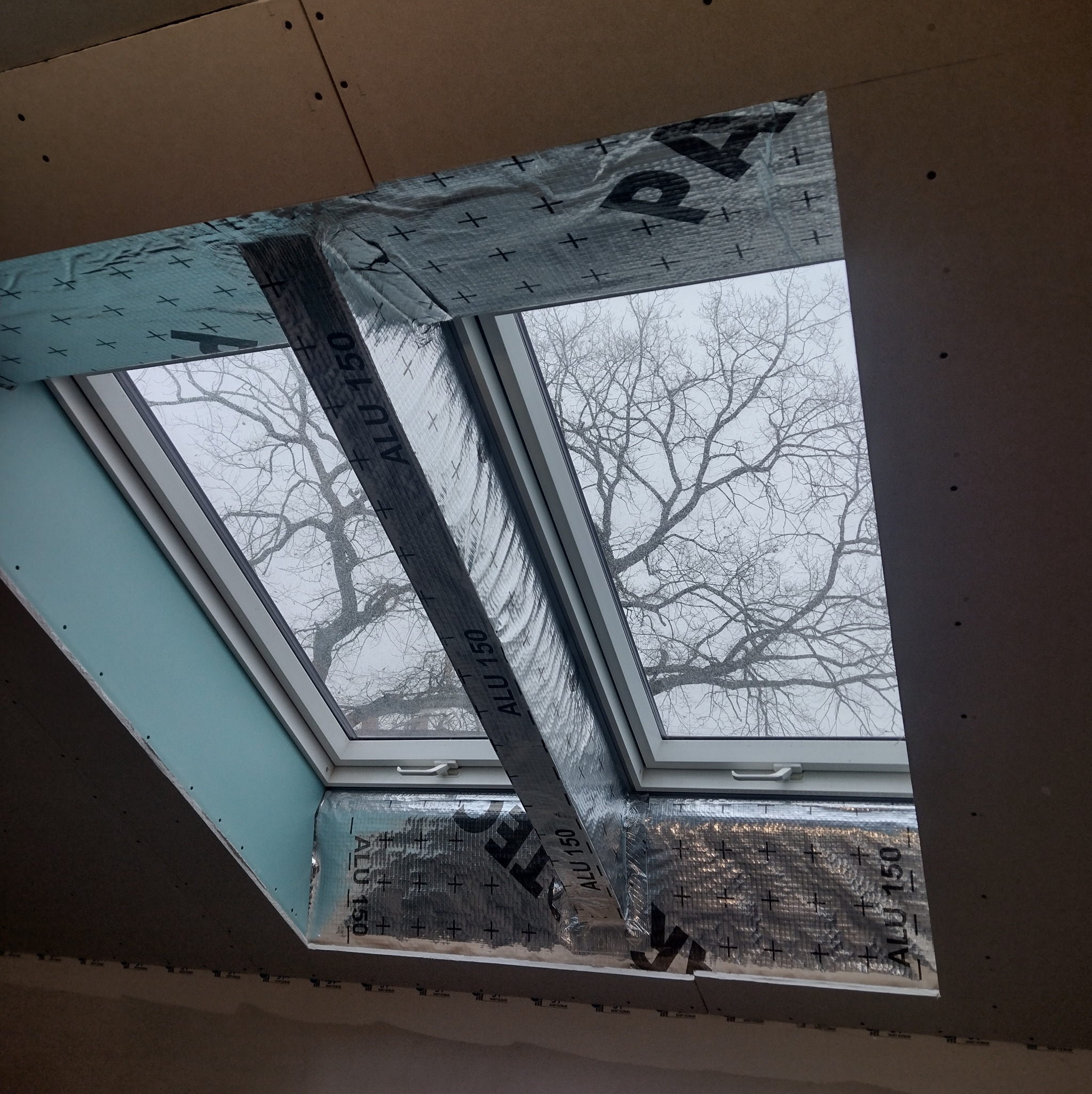 Montaż okna dachowego z izolacją ALU 150, widok na drzewo przez szybę. Płyta karton-gips na poddaszu w trakcie prac wykończeniowych. Ujęcie z dołu.