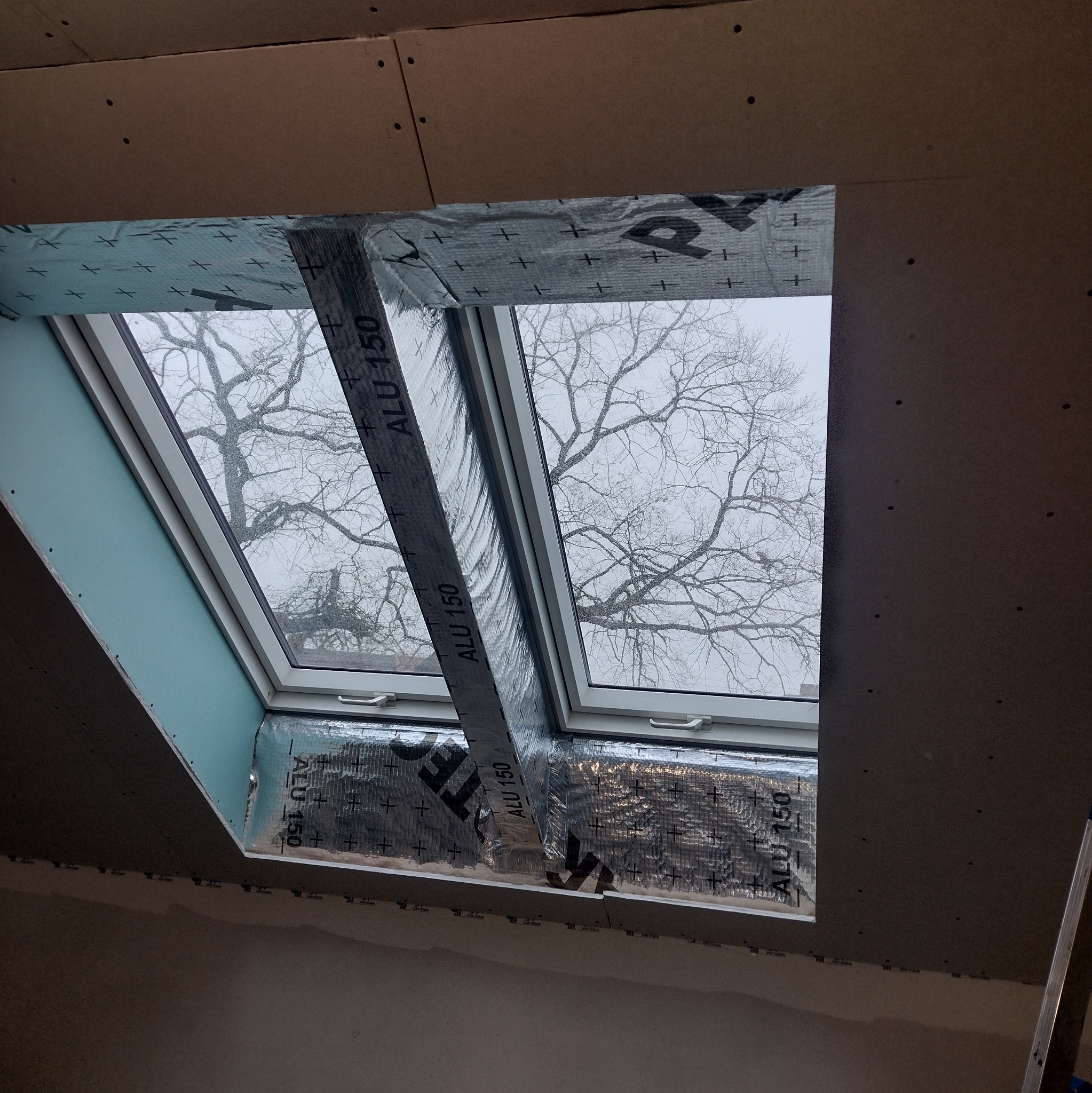 Montaż okna dachowego z izolacją i płytami gipsowo-kartonowymi. Widok na drzewo przez okno. Etap prac wykończeniowych.