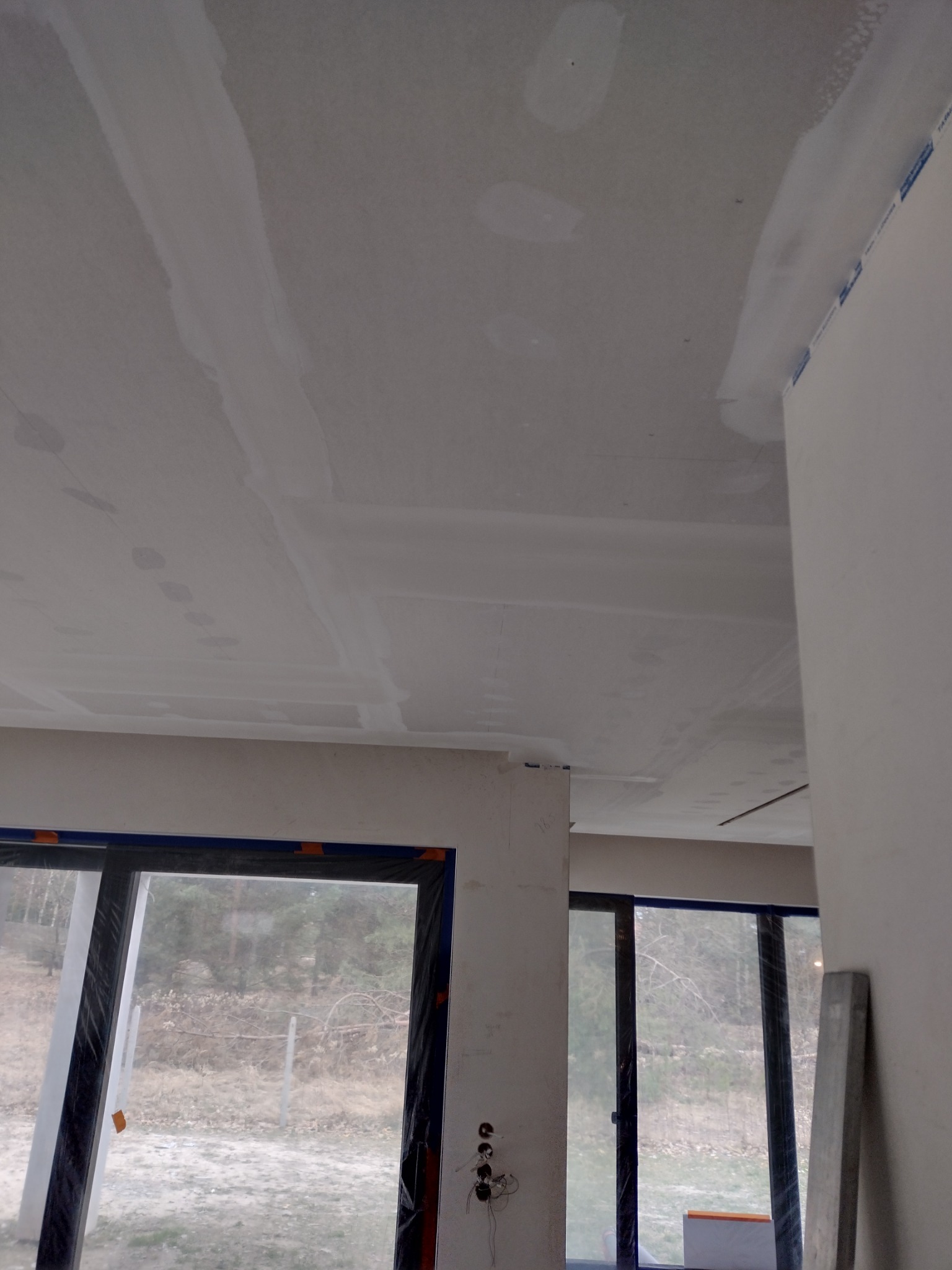 Płyty karton-gips na suficie i ścianach, widoczne łączenia szpachlowane. Okna zabezpieczone folią. W tle widok na zewnątrz.