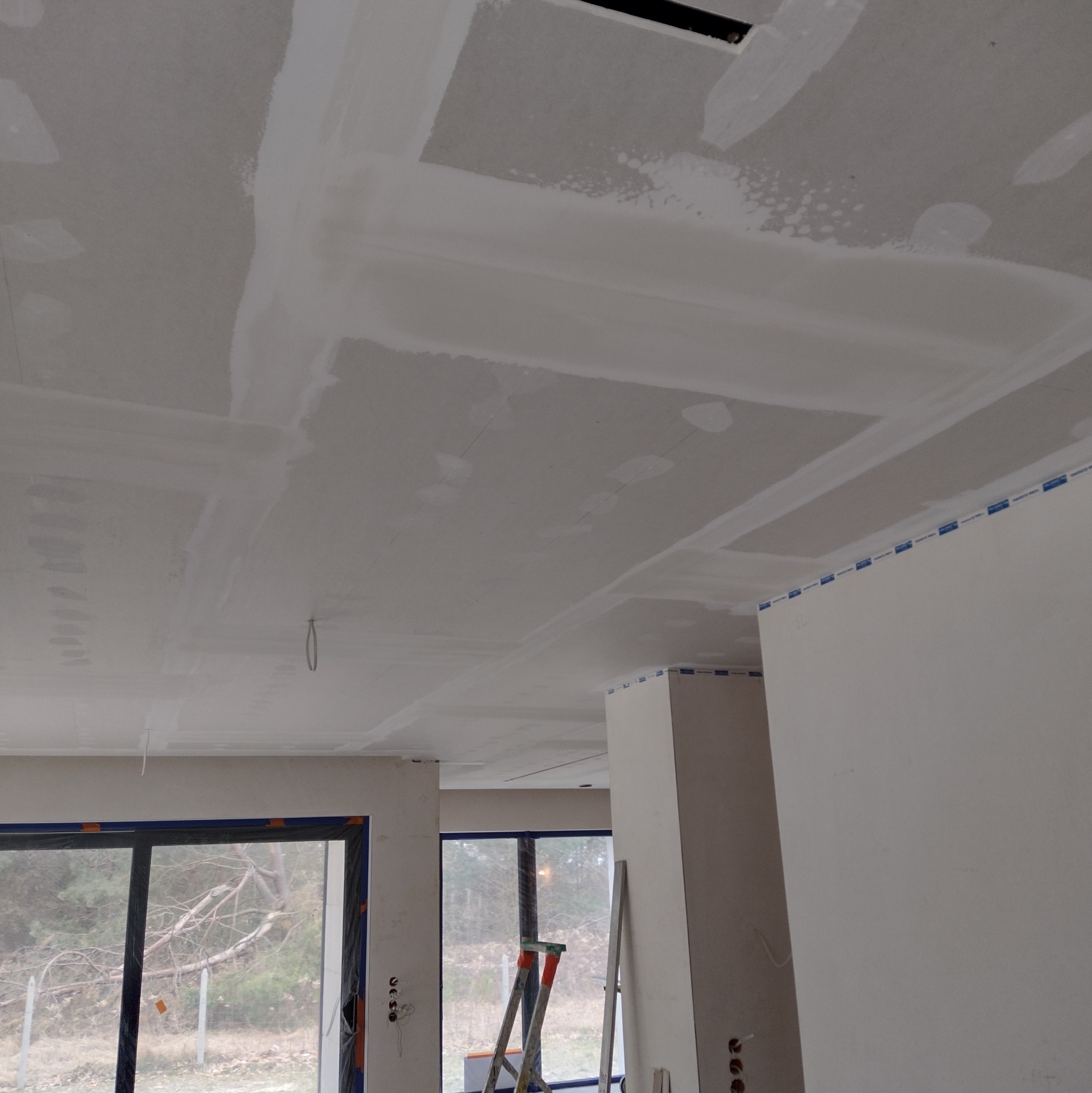 Sufit z płyt kartonowo-gipsowych po szpachlowaniu, widoczne łączenia i otwory wentylacyjne. W tle okno i drabina malarska.