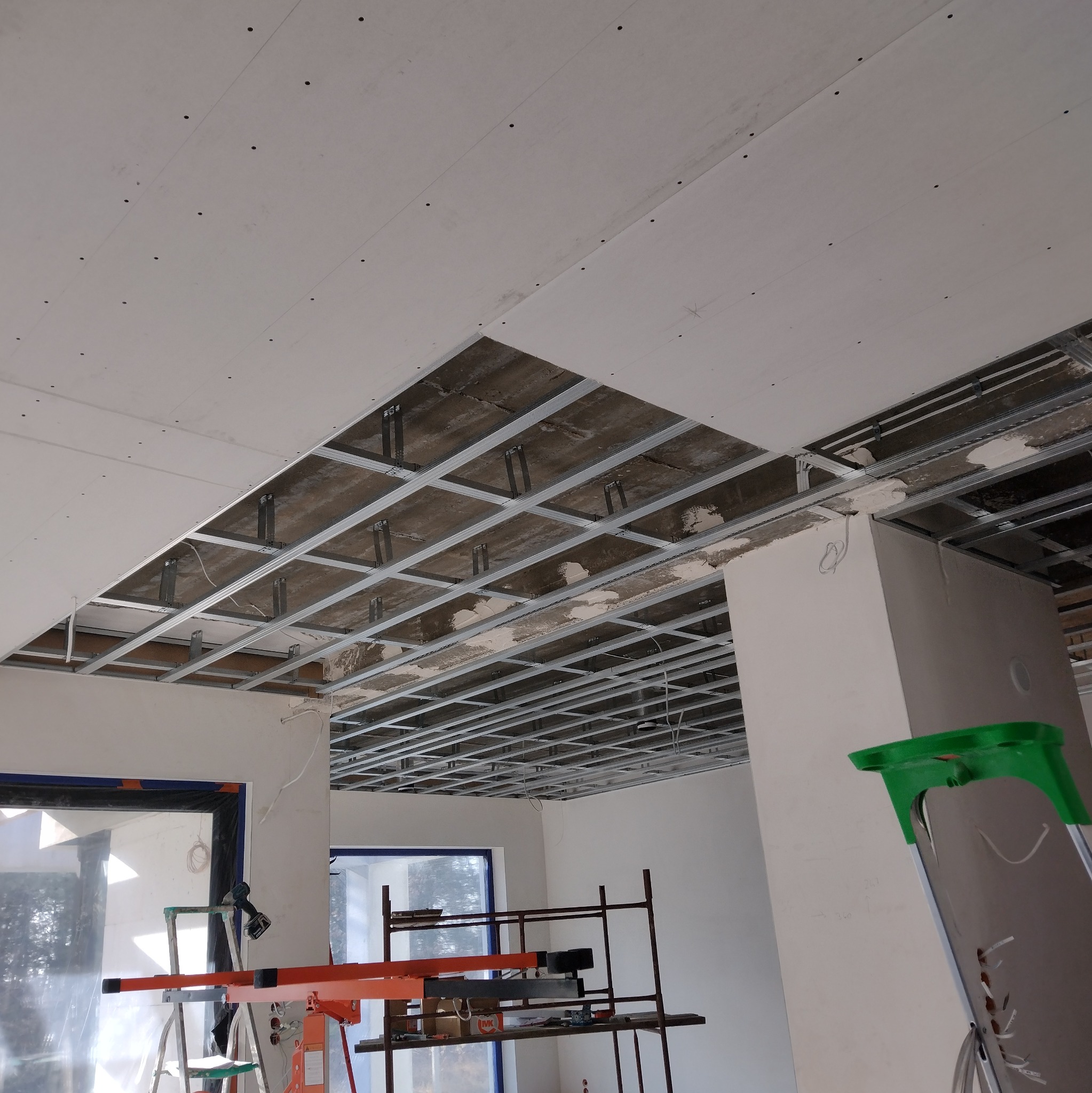 Montaż sufitu podwieszanego z płyt karton-gips na metalowej konstrukcji. Widoczne profile i częściowo zamontowane płyty. W tle okna i drabina.
