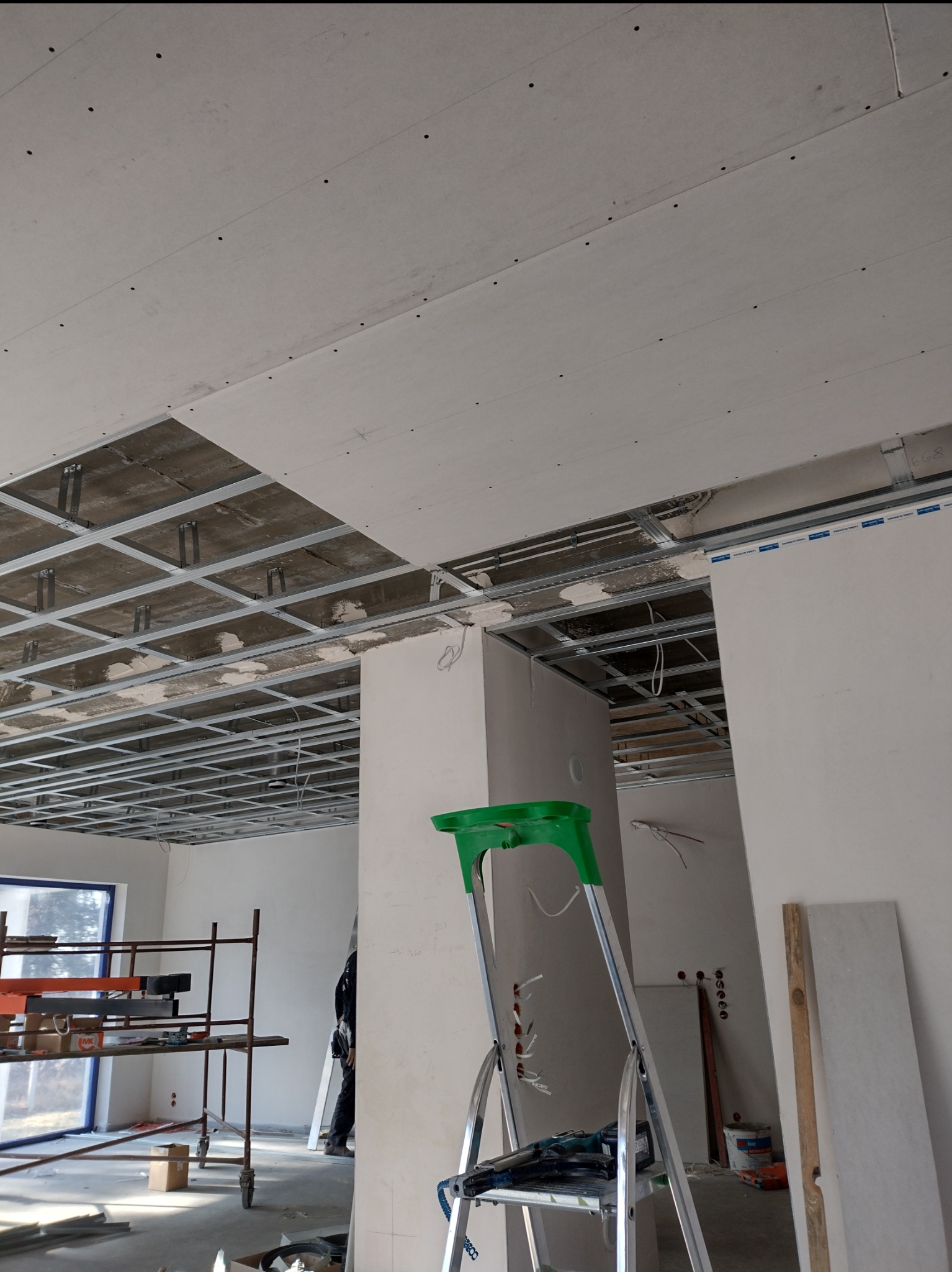 Sufit podwieszany z płyt gipsowo-kartonowych w trakcie montażu, widoczna metalowa konstrukcja, drabina i rusztowanie w pomieszczeniu. Etap prac wykończeniowych.