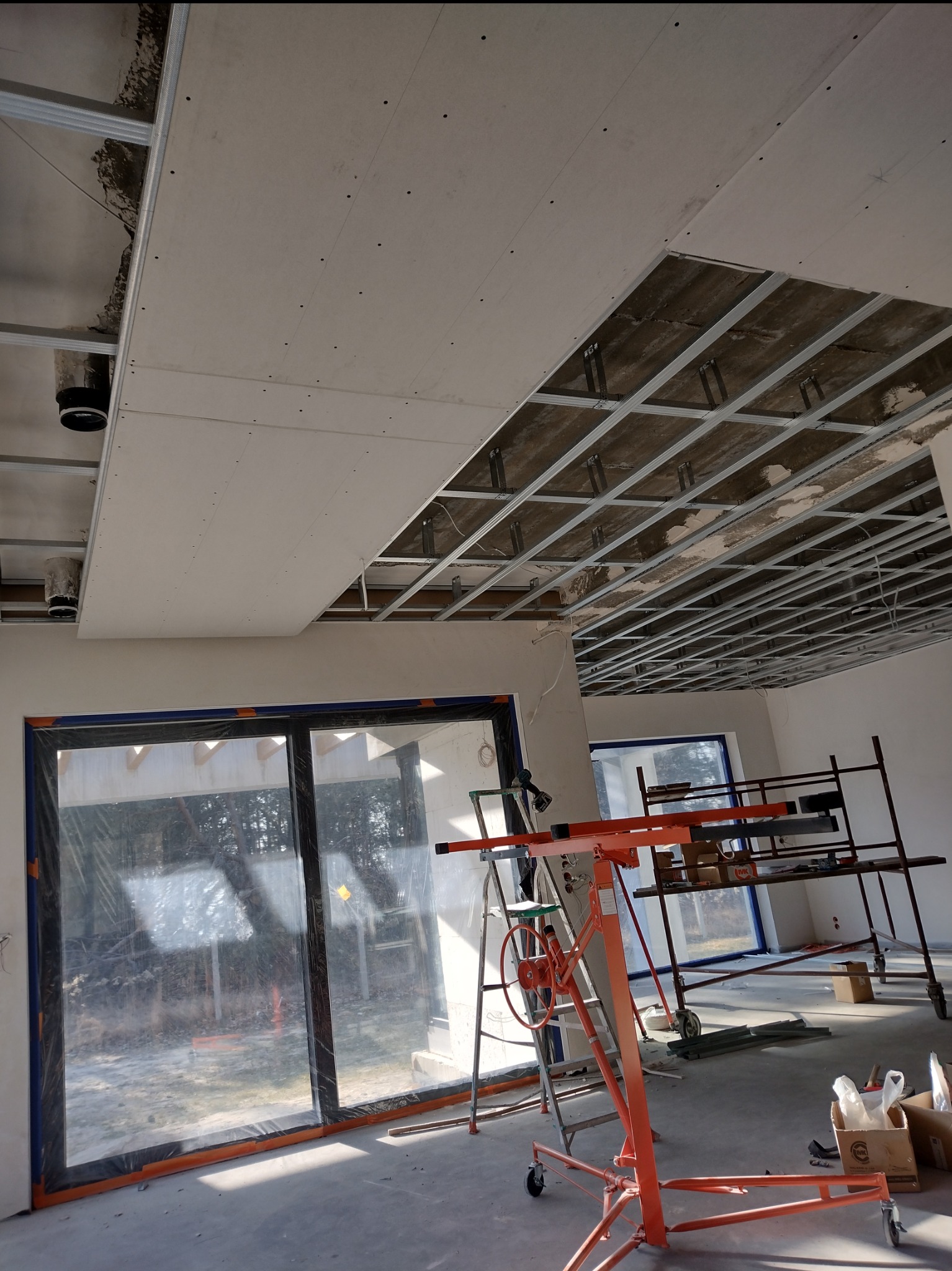 Montaż sufitu podwieszanego z płyty gipsowej, widoczny stelaż i podnośnik. Okna zabezpieczone folią malarską. Częściowo zamontowane płyty.