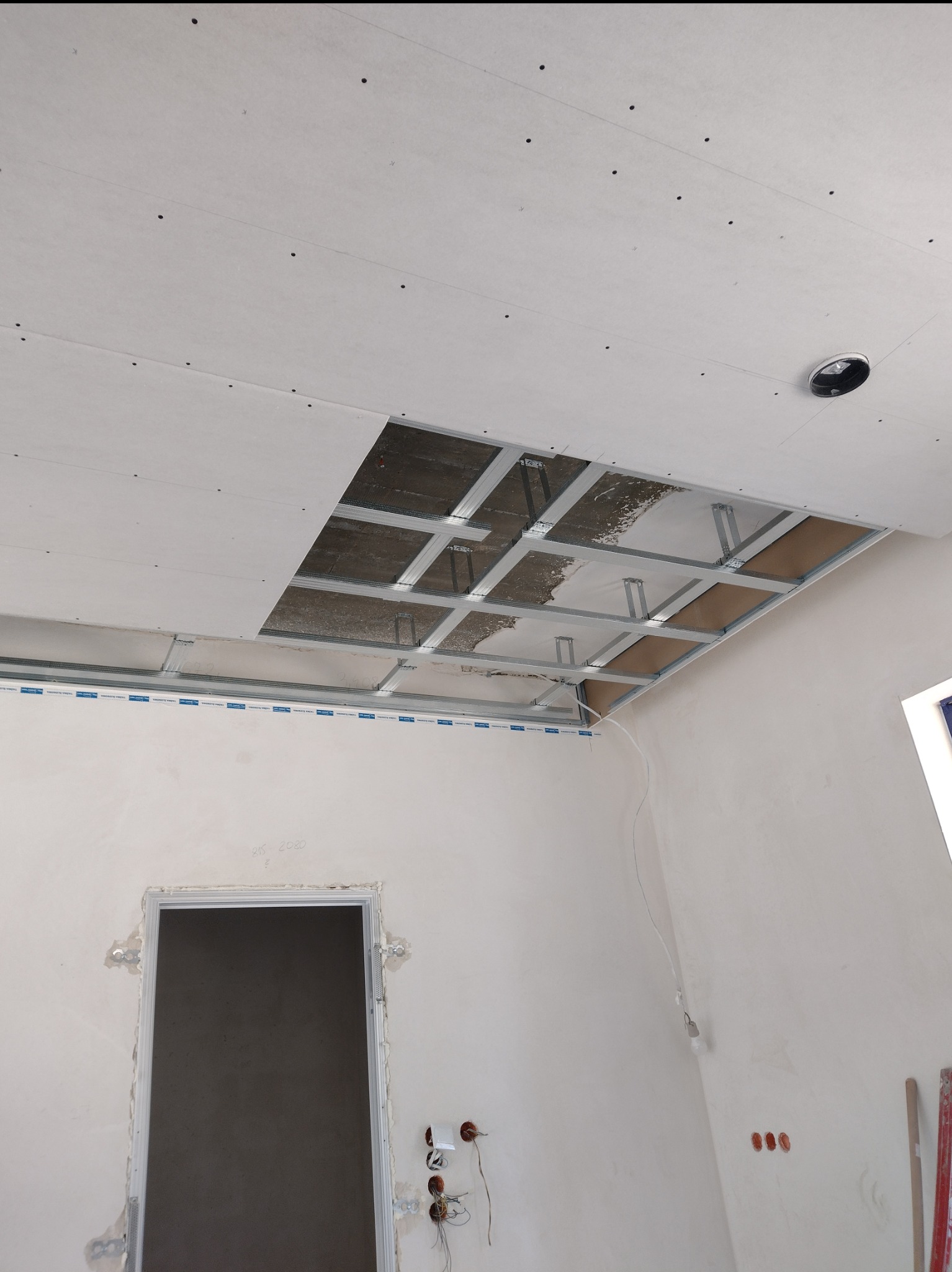 Montaż sufitu podwieszanego z płyt karton-gips: widoczne metalowe profile i częściowo zamontowane płyty. W tle ściany w trakcie prac wykończeniowych.