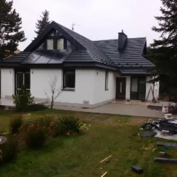 Nowoczesny dom jednorodzinny z białymi ścianami i grafitowym dachem, widoczne pozostałości po pracach dekarskich na trawniku, w tle iglaki.