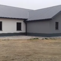 Nowo wybudowany, parterowy budynek z dachem z blachy trapezowej, pomalowany na biało i szaro, z ciemnymi oknami i drzwiami, na niezagospodarowanej działce.