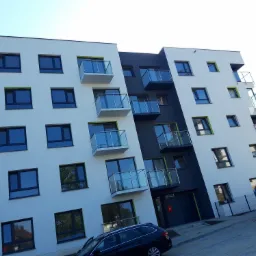 Nowoczesny budynek wielorodzinny z białymi i grafitowymi elewacjami, balkonami z szklanymi balustradami i oknami z zielonymi akcentami, widziany z dołu, z zaparkowanym samochodem osobowym...