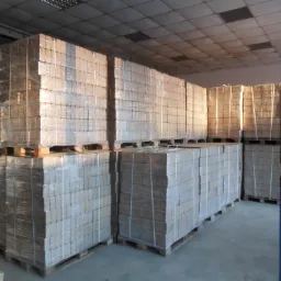 Magazyn wypełniony paletami z brykietem drzewnym owiniętym folią, widoczne oświetlenie sufitowe i słup podtrzymujący.