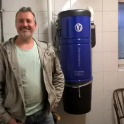 Uśmiechnięty mężczyzna w jasnej kurtce i jeansach stoi obok niebieskiego odkurzacza centralnego Variovac Premium 240 VIP zamontowanego na kafelkowanej ścianie w piwnicy.