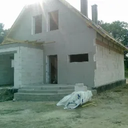 Surowy stan domu murowanego z betonu komórkowego, widoczne otwory okienne i drzwiowe, schody wejściowe i przygotowane materiały budowlane przed budynkiem.