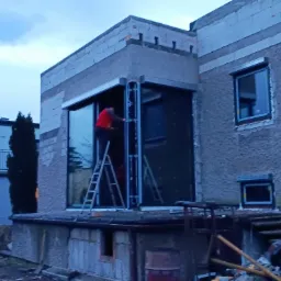 Montaż drzwi przesuwnych w surowym stanie budynku, pracownik na drabinie w czerwonej koszulce, widoczne okna z roletami zewnętrznymi.