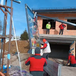 Montaż dużej szyby okiennej za pomocą dźwigu na budowie, widoczni pracownicy w odblaskowych ubraniach, surowe otoczenie budowy z elementami śniegu.