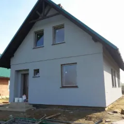 Dom jednorodzinny po tynkowaniu elewacji na szaro, z widocznymi oknami z brązowymi ramami i zielonym dachem. Na ziemi leżą rury.