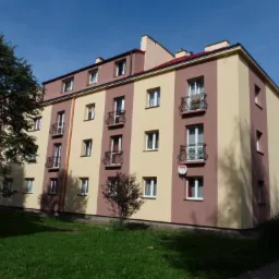 Odnowiona elewacja bloku z balkonami i nową anteną satelitarną, pomalowana w pionowe pasy beżu i brązu, na tle zielonego trawnika i błękitnego nieba.