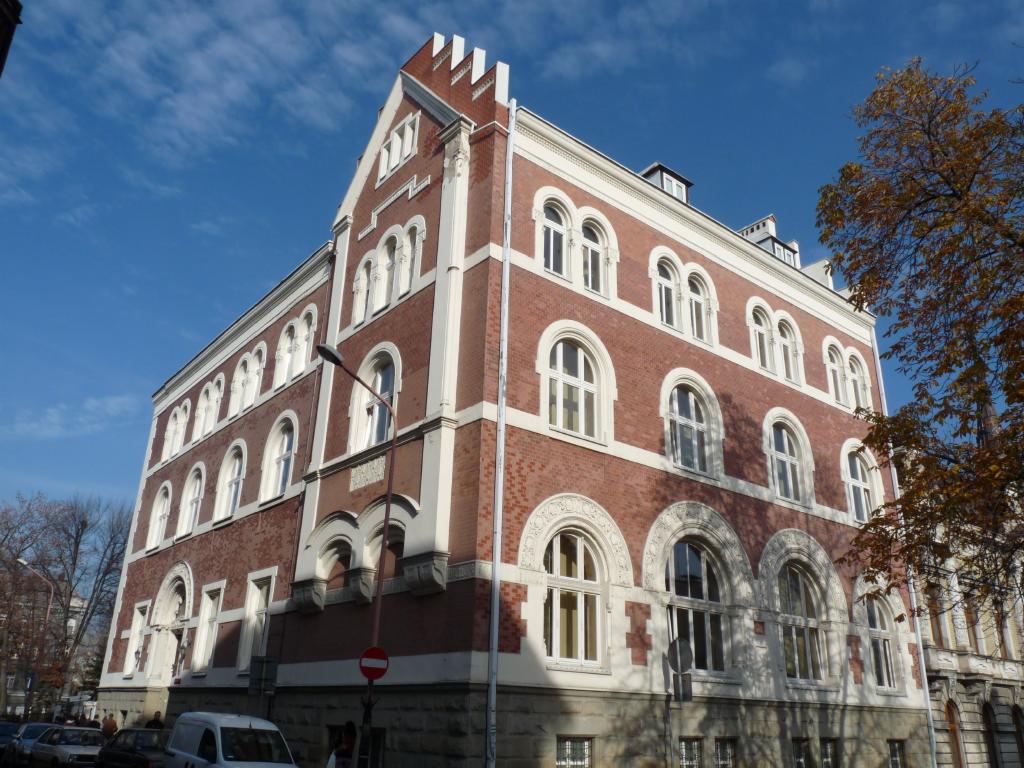 Elewacja odrestaurowanego, ceglanego budynku z białymi elementami dekoracyjnymi i łukowymi oknami. Widok na fasadę w jesiennym słońcu.