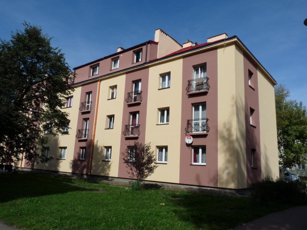 Odnowiona elewacja bloku z balkonami i nową anteną satelitarną, pomalowana w pionowe pasy beżu i brązu, na tle zielonego trawnika i błękitnego nieba.