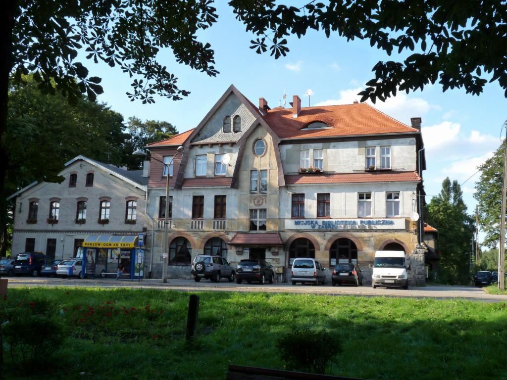 Budynek Miejskiej Biblioteki Publicznej w Zabrzu-Filii nr 5. Dwupiętrowa konstrukcja z czerwonym dachem i elementami architektonicznymi. Widok z ulicy w słoneczny dzień.