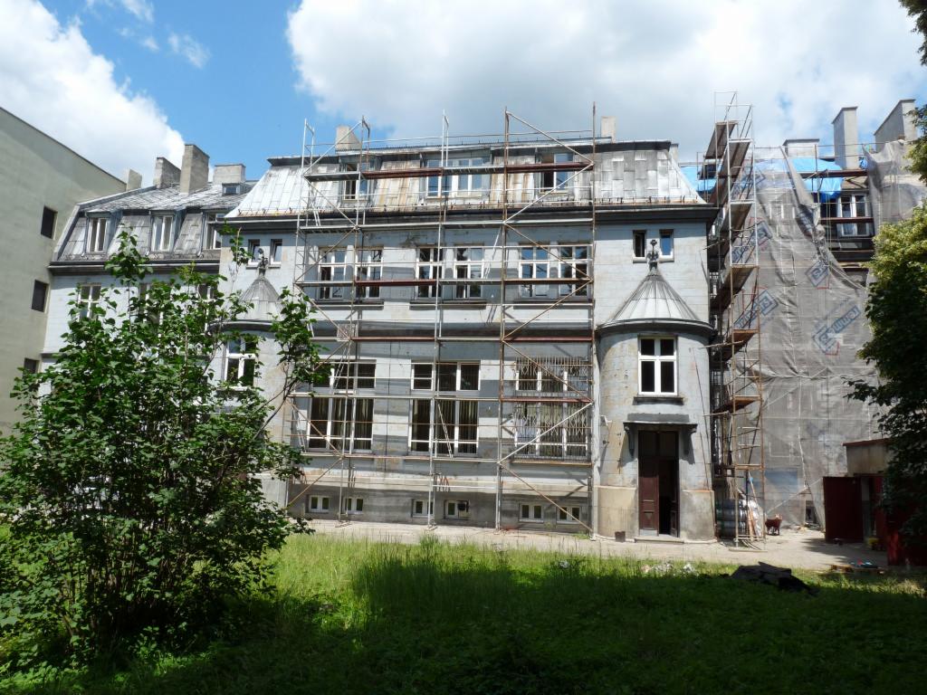 Renowacja kamienicy z rusztowaniami, częściowo zakryta plandeką. Widoczna zieleń i błękitne niebo. Prace konserwatorskie w toku.