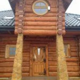 Drewniany dom z bali z okrągłym oknem i dekoracyjnymi drzwiami wejściowymi, widok z dołu, podkreślający naturalną fakturę drewna i solidną konstrukcję budynku.