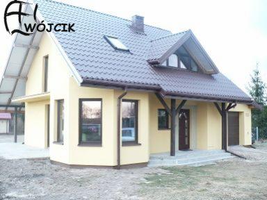 Żółty dom jednorodzinny z brązowym dachem i stolarką okienną, widoczny z perspektywy trawnika. Charakterystyczny daszek nad wejściem wsparty na drewnianych słupach.