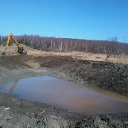 Żółta koparka przy wykopanym stawie wypełnionym wodą. Widoczne świeże ślady ziemi, w tle las i błękitne niebo. Krajobraz po robotach ziemnych.