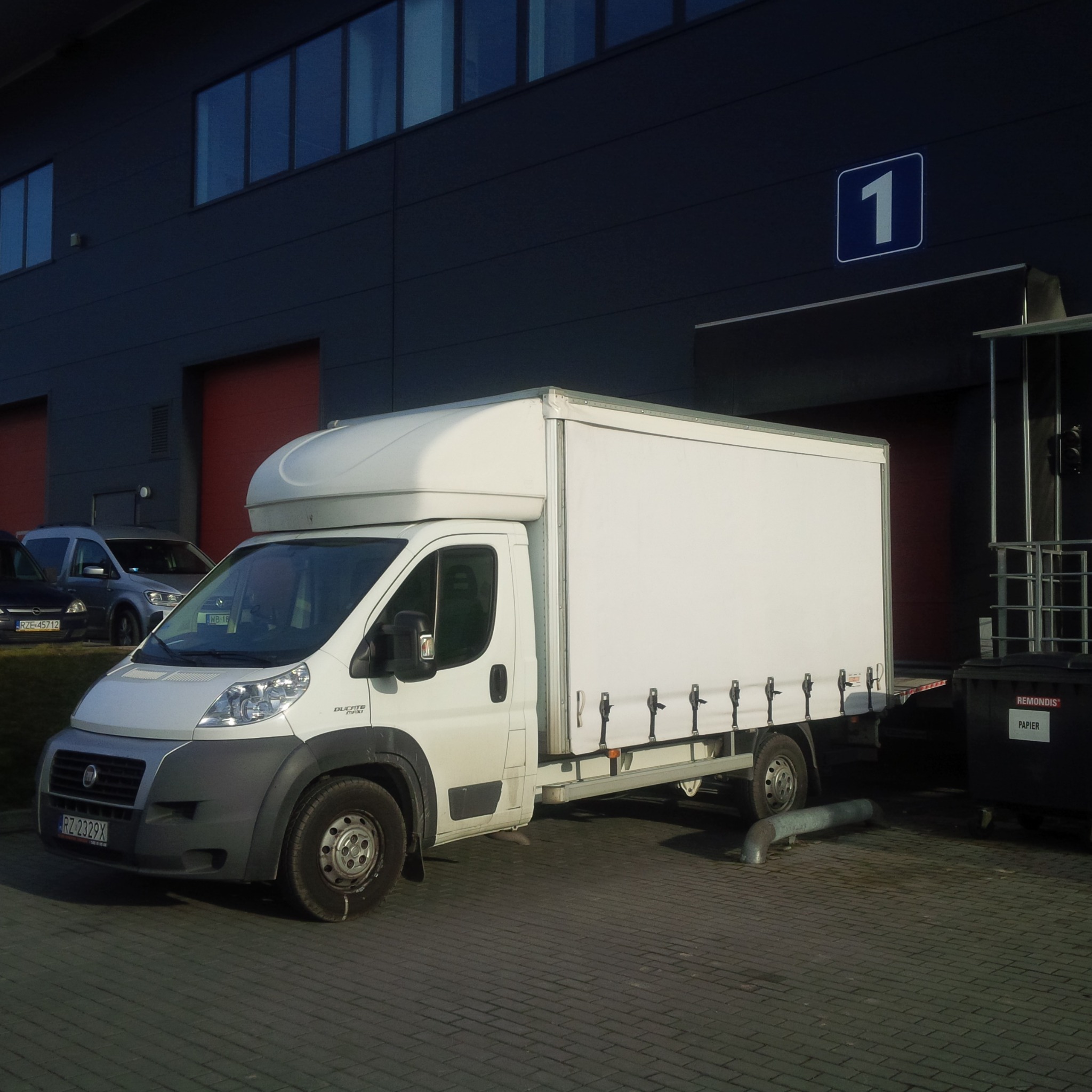 Biały samochód dostawczy Fiat Ducato Maxi z plandeką na tle szarego budynku z numerem 1 i czerwonymi bramami, zaparkowany na brukowanym placu.