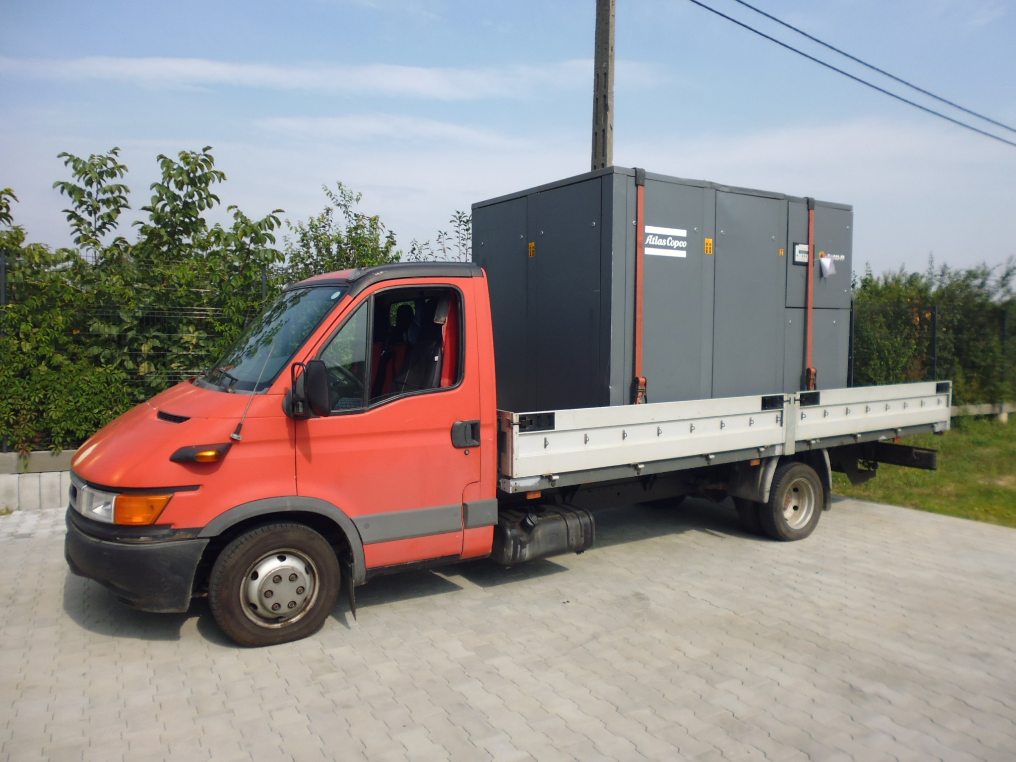 Czerwony samochód dostawczy z platformą transportową przewożący duży, szary agregat Atlas Copco na tle zieleni i błękitnego nieba.
