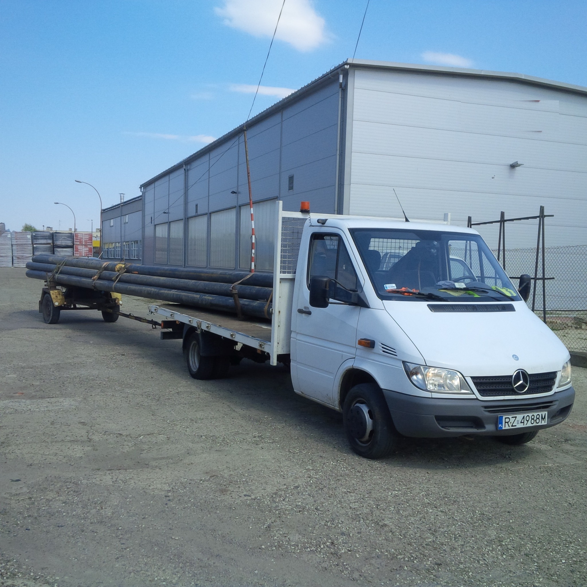 Biały samochód dostawczy Mercedes-Benz z przyczepą przewożący rury na tle hali magazynowej w Rzeszowie.