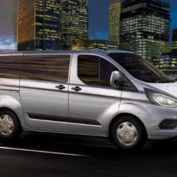 Srebrny Ford Transit Custom przewożący osoby na tle miejskiego krajobrazu z wieżowcami oświetlonymi nocą.