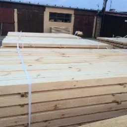 Stosy jasnej tarcicy budowlanej, spięte białymi taśmami, na tle magazynu z ciemnymi bramami i oknem. Widoczne sęki i naturalny rysunek drewna.