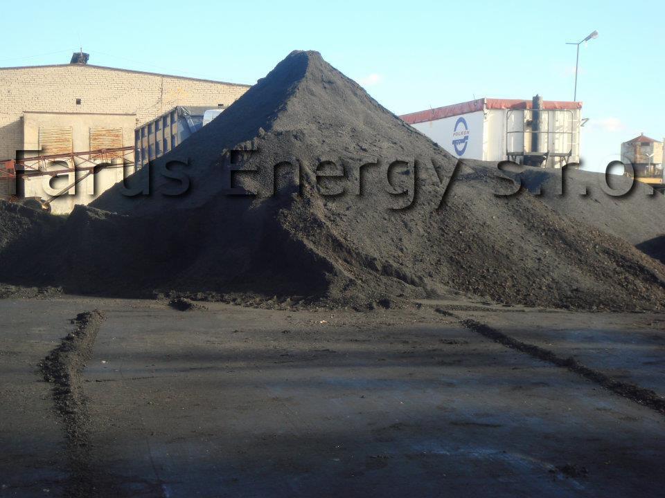 Góra miału węglowego na placu składowym, widoczne ślady opon pojazdu, w tle budynek magazynowy i naczepa ciężarówki z logo.