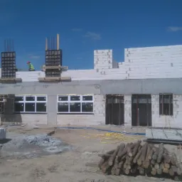 Wznoszenie ścian budynku z bloczków betonu komórkowego, widoczne zbrojenia i szalunki, na budowie pracownik w kamizelce odblaskowej, w tle błękitne niebo.