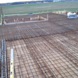 Zbrojenie fundamentu z siatki stalowej na drewnianym szalunku, widoczny poziomica i wystające pręty pionowe.