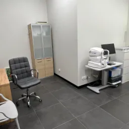 Wnętrze gabinetu lekarskiego z biurkiem, krzesłem biurowym, szafką i aparaturą diagnostyczną do badania wzroku (autorefraktometr) na stoliku z monitorem.