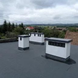 Widok na płaski dach pokryty papą termozgrzewalną z trzema kominami wentylacyjnymi na tle zabudowy jednorodzinnej i pochmurnego nieba.